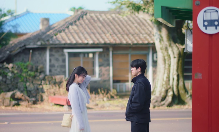 Yumi e Yoo Babi frente a frente em um ponto de ônibus de rua, em Jeju