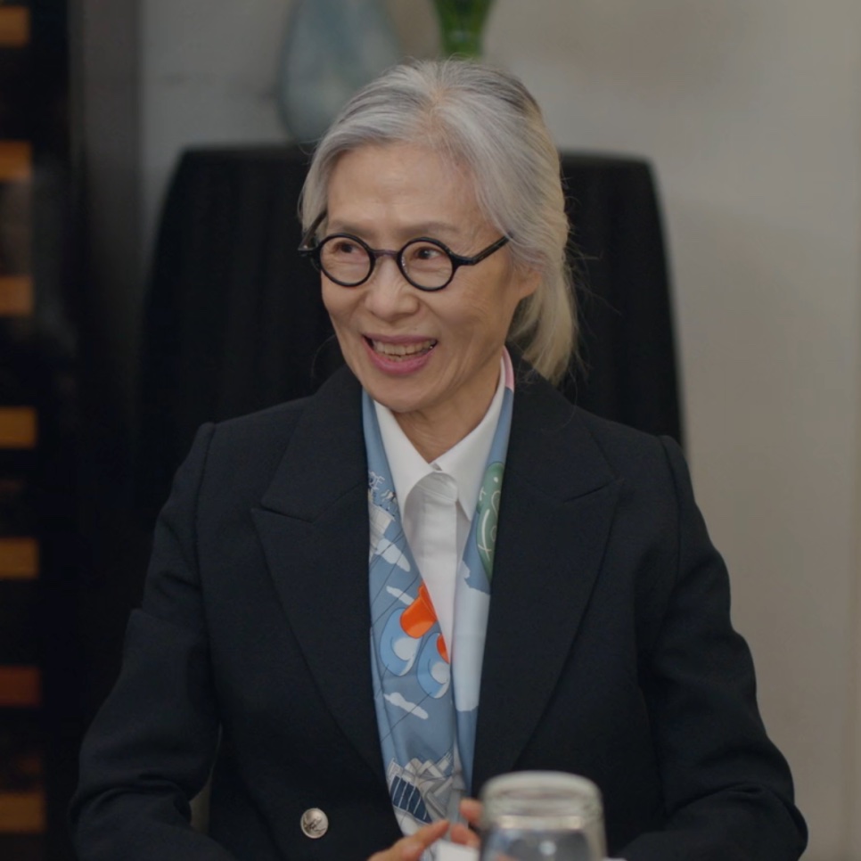 Baek Kyung-ja, a presidente do grupo Reina, de terno preto e óculos redondos, falando numa reunião