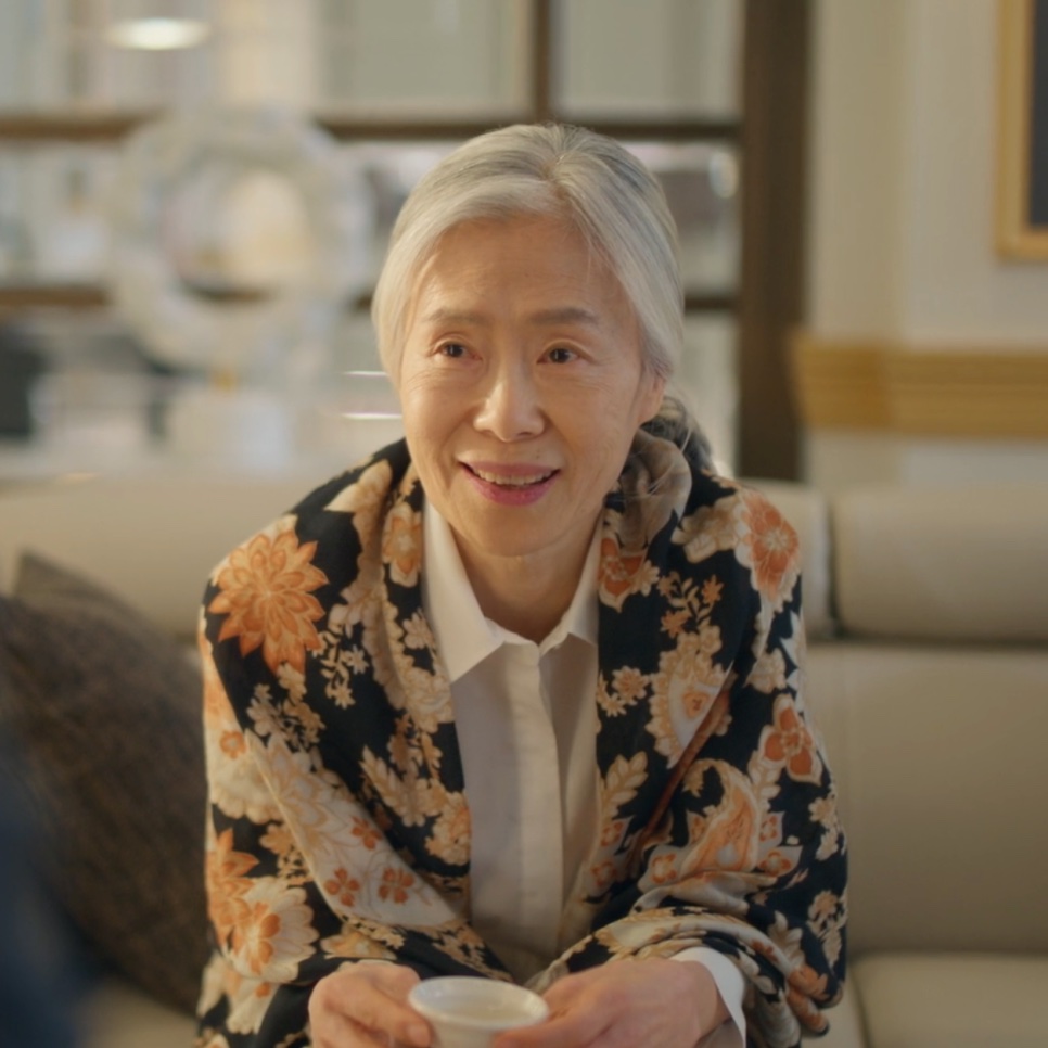 Baek Kyung-ja de casaco florido, sentada na sala de casa com uma xícara nas mãos