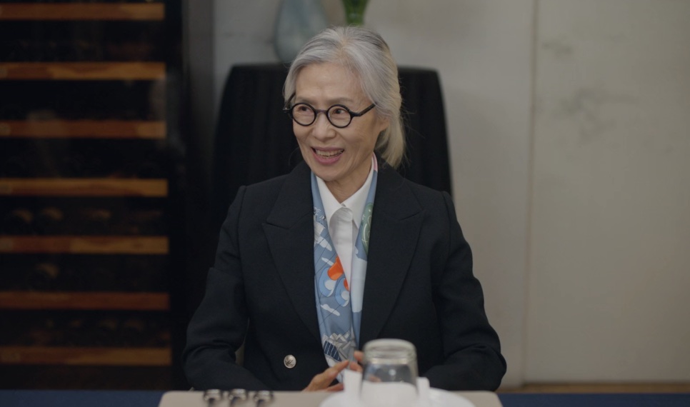 Baek Kyung-ja sentada à mesa de uma reunião, de terno preto e lenço no pescoço
