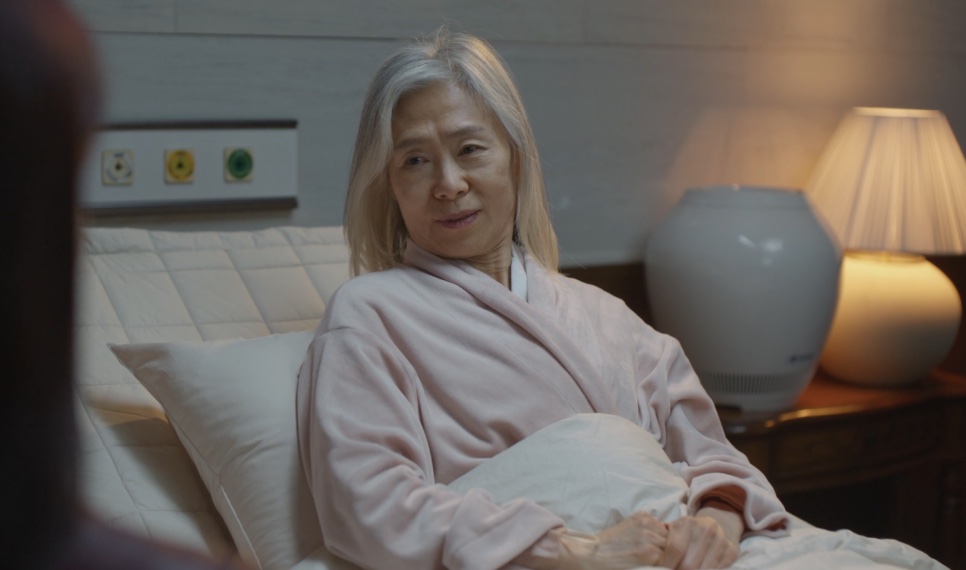 Baek Kyung-ja deitada numa cama de hospital, de roupão rosa, com um abajur aceso ao lado