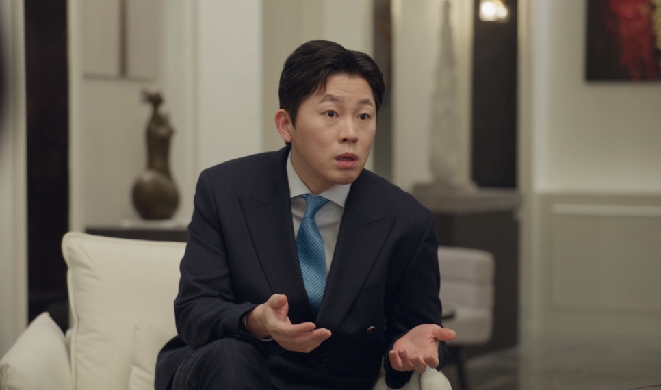 Cheon Dong-jun, primo mais velho da Yeo-ri, de terno, falando com as mãos abertas no sofá