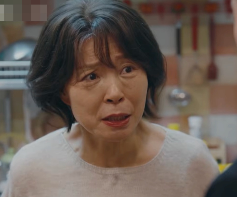 Ok Seon-ja de blusa clara, na cozinha, falando com alguém