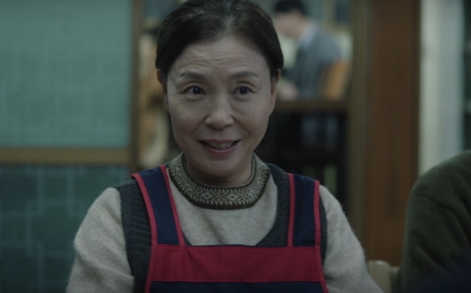 Yu Jung-won de avental, sorrindo dentro do restaurante dela