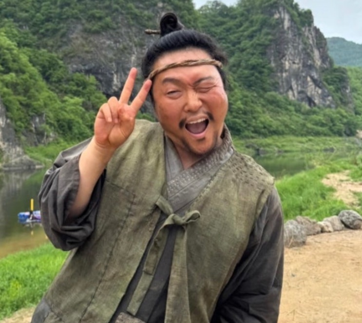 Lee Jun-hyuk de hanbok e faixa na cabeça, fazendo sinal de V numa paisagem de montanha