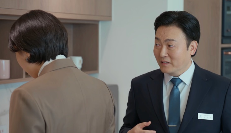 Bong Ji-uk sentado à mesa conversando com outro homem