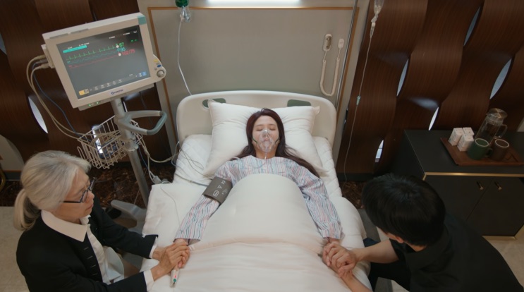 Yeo-ri deitada numa cama de hospital, com monitores ao lado e duas pessoas ao redor