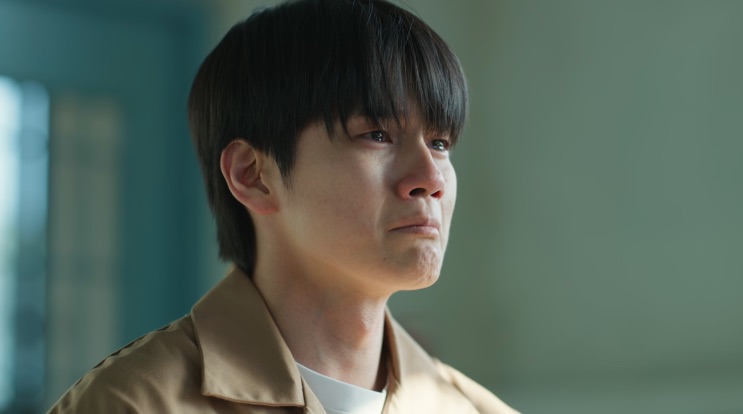 Kang Min-hwan de casaco claro, olhando para o lado com expressão dura