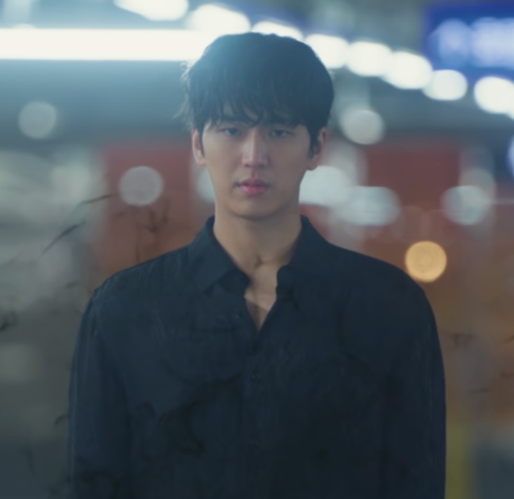 Ji-hwan de camisa preta numa rua à noite, com as luzes desfocadas atrás