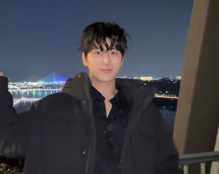 Selfie do ator Kim Min-chul, que faz o Ji-hwan, de jaqueta preta à noite, com um rio iluminado atrás