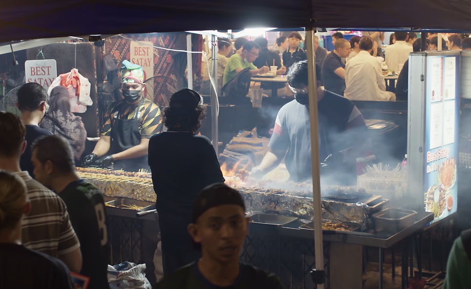 Barracas de espetinhos na brasa, com fumaça e pessoas em volta
