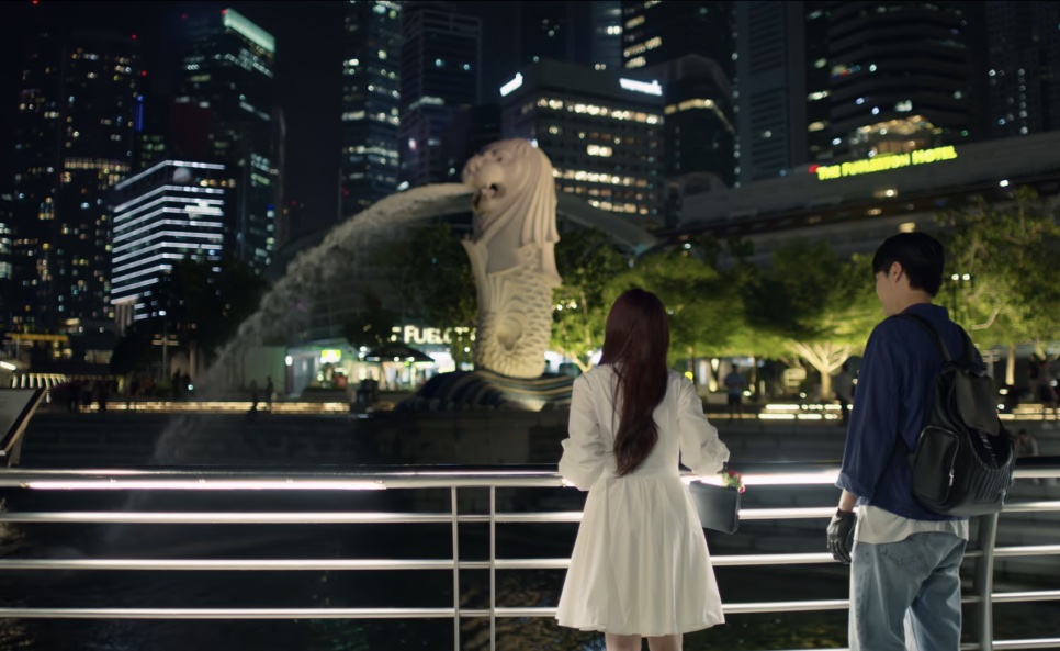 Yeo-ri e Gang-uk de costas, olhando o Merlion à noite