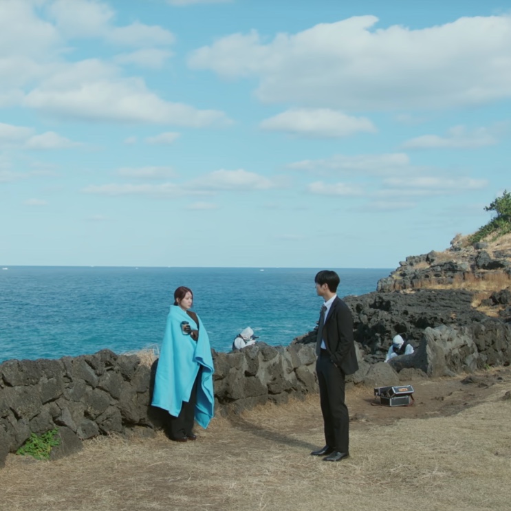 Yeo-ri enrolada num cobertor azul e Kang-wook diante do mar, no penhasco de Jeju