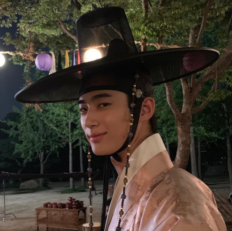 Byeon Woo-seok de hanbok e chapéu tradicional, à noite, com lanternas ao fundo
