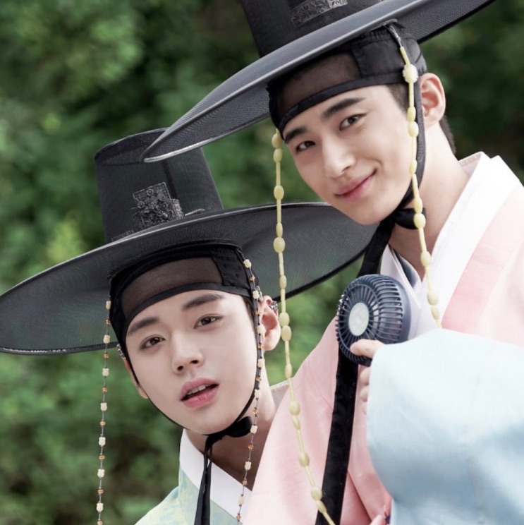 Byeon Woo-seok e Park Ji-hoon de hanbok e chapéu tradicional, lado a lado, sorrindo para a câmera
