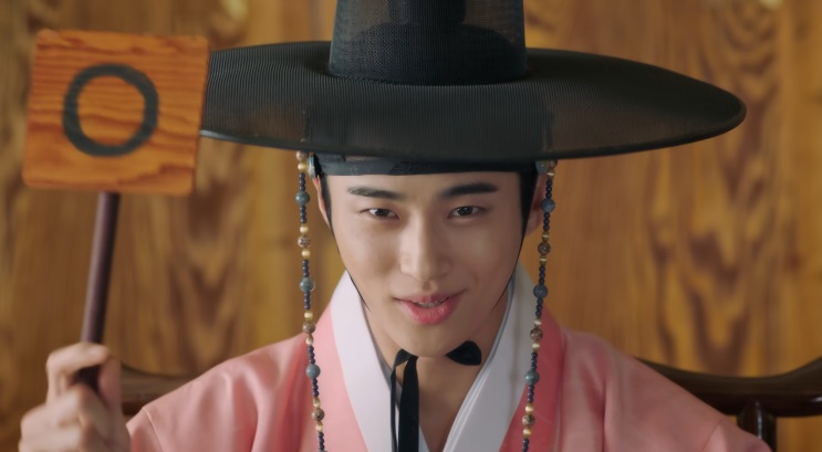 Byeon Woo-seok de hanbok rosa e chapéu, segurando uma placa de madeira