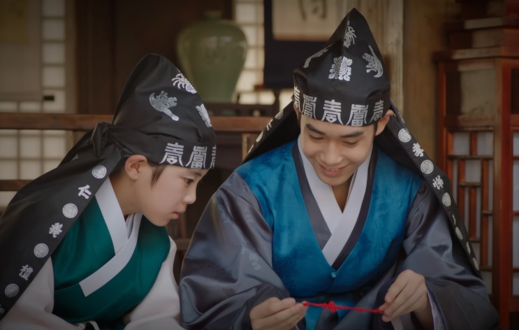 Um rapaz de hanbok azul e capuz de casamenteiro sorrindo enquanto mexe em um cordão vermelho, ao lado de um menino