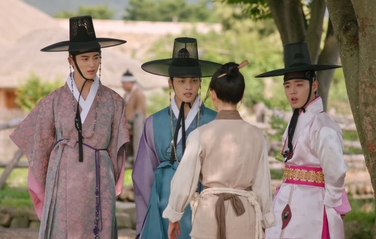 Três casamenteiros de hanbok colorido e chapéu de aba larga conversando com uma mulher na rua