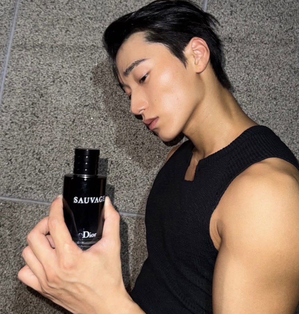 Choi San deitado, de regata preta, segurando um frasco de perfume
