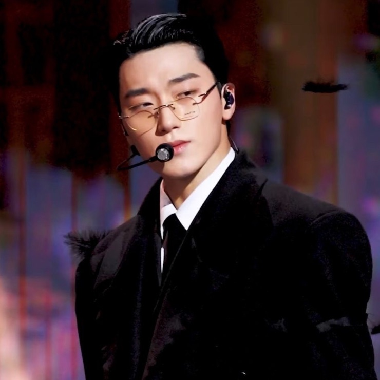 Choi San do ATEEZ no palco, de terno preto e óculos de armação fina, ar imponente