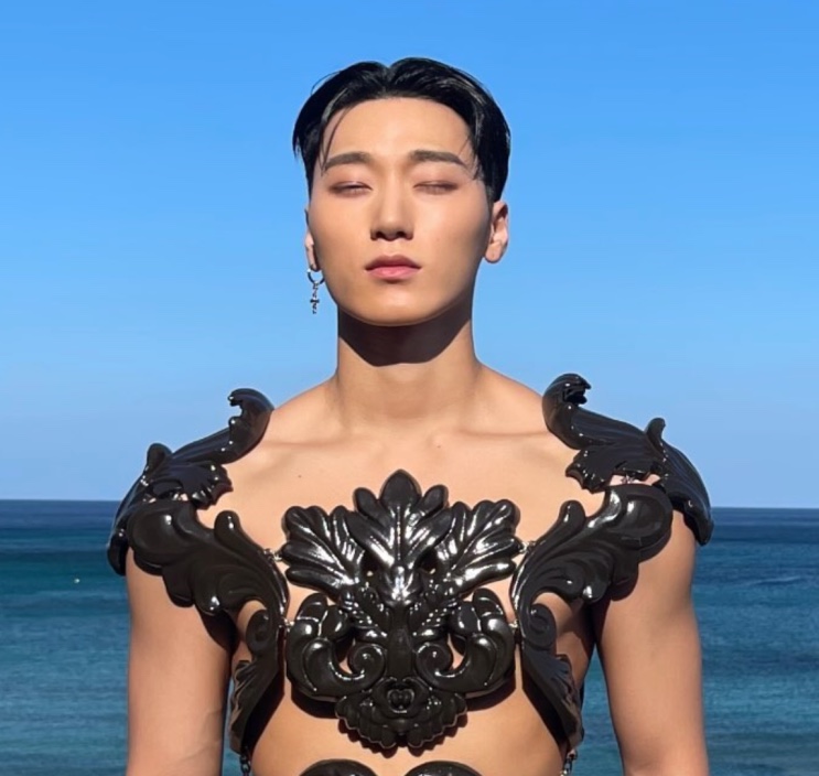 Choi San com o mesmo peitoral preto, de olhos fechados, tendo o mar azul ao fundo