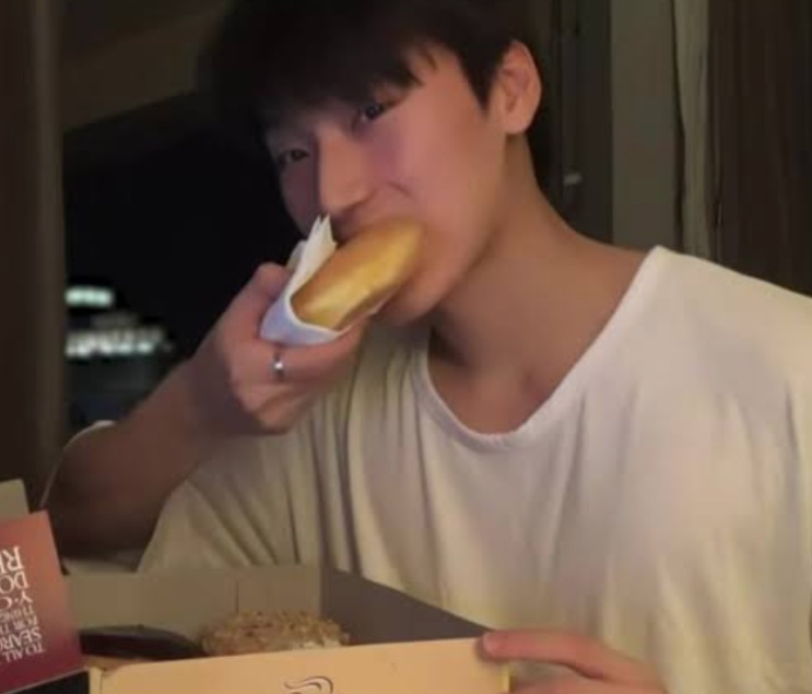 Choi San de camiseta branca dando uma mordida num pão, sentado à mesa