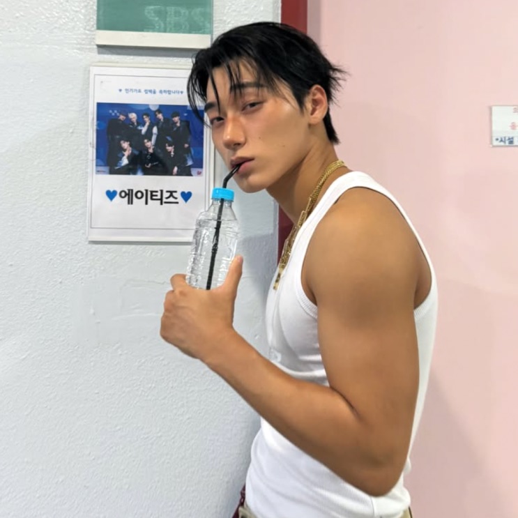 Choi San do ATEEZ de regata, com pôster do grupo atrás