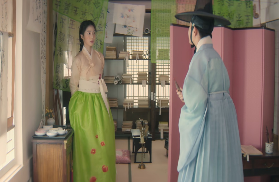 A jovem Cho Yun, de saia verde, e o jovem Cho Won, de chapéu gat e roupa azul-clara, numa sala cheia de livros, em Escândalo e Intriga