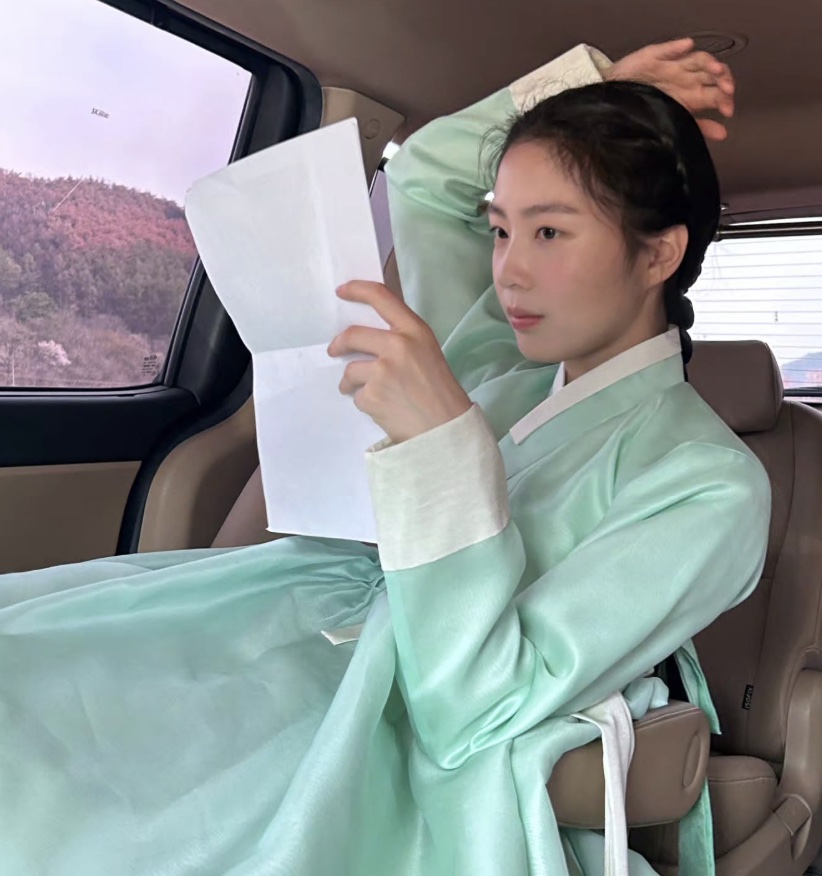 Shin So-hyun de hanbok verde-claro, com um papel na mão, sentada dentro de um carro