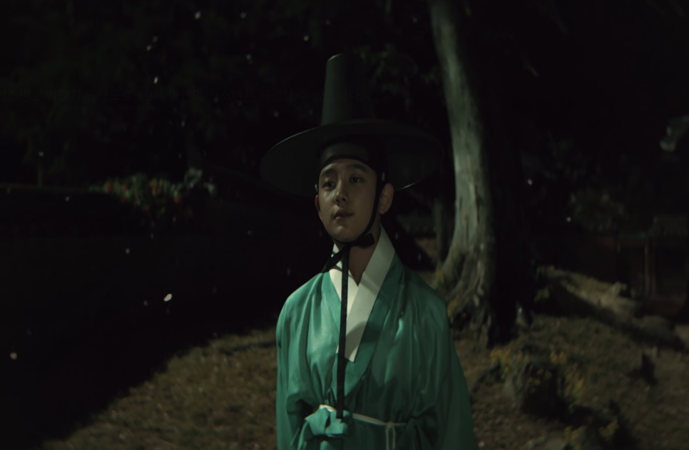 O jovem Cho Won, de roupa verde e chapéu gat, parado num jardim à noite, em Escândalo e Intriga