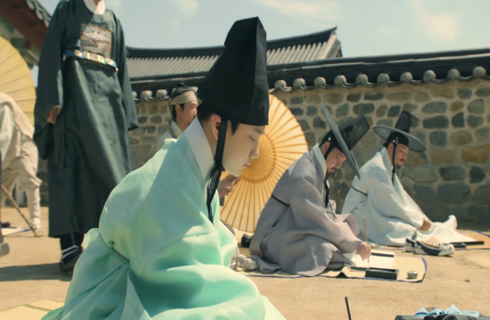 O jovem Cho Won, de roupa verde-clara e chapéu gat, sentado no chão de um pátio durante um exame, com outros candidatos ao fundo, em Escândalo e Intriga