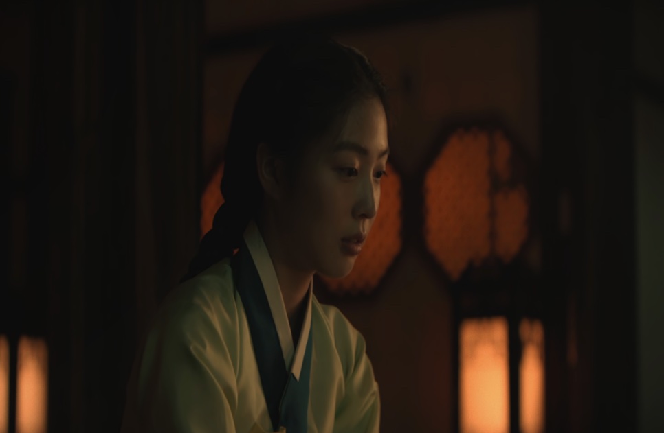 A jovem Cho Yun, de hanbok claro, olhando para baixo num quarto iluminado por lanternas, em Escândalo e Intriga