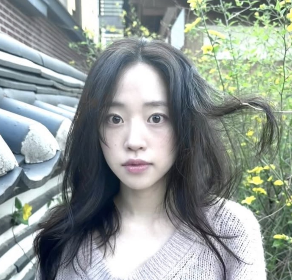 Kim Ji-an de suéter claro, com o cabelo ao vento, diante de um telhado de telhas e um arbusto florido de amarelo