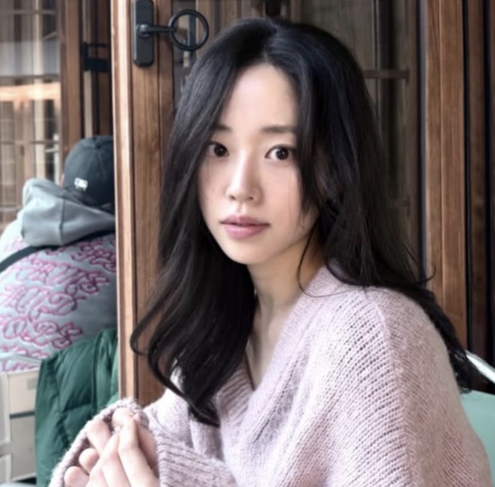 Kim Ji-an de tricô rosa claro, apoiada numa porta de madeira, olhando para a câmera