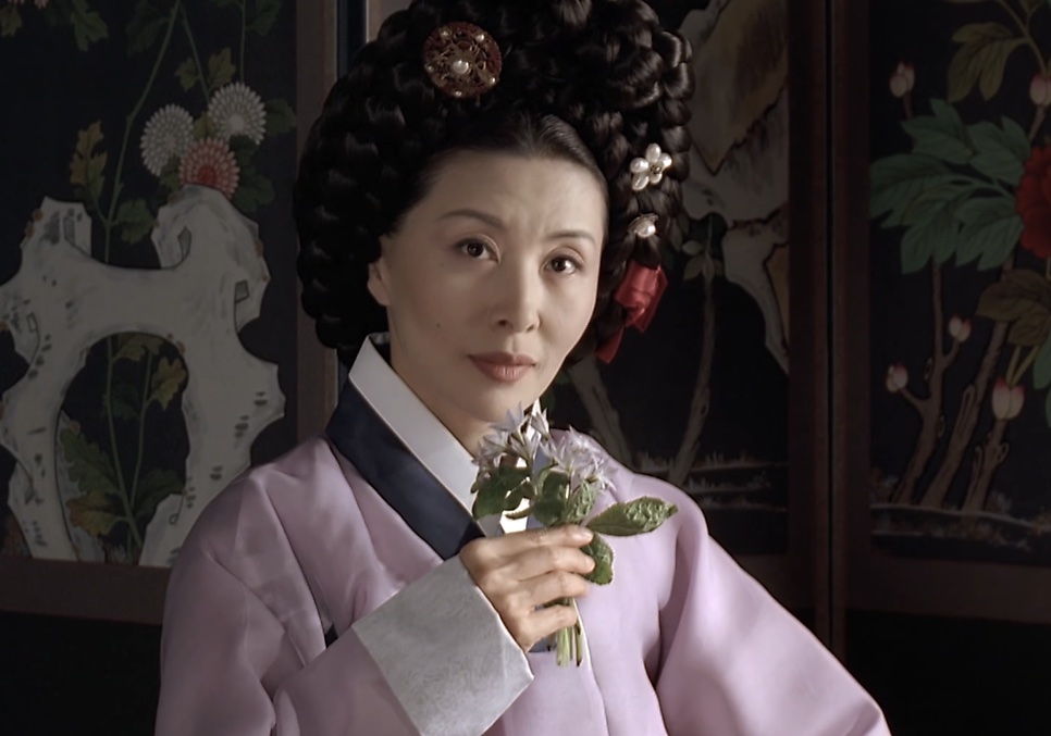 A Senhora Jo, de hanbok lilás, segurando um raminho de flores diante de um biombo pintado