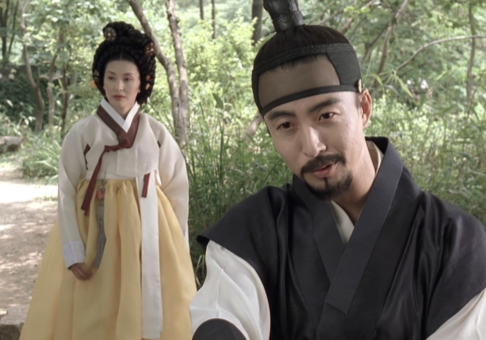 O Jo-won sorrindo numa trilha no meio das árvores, com uma mulher de hanbok amarelo mais atrás