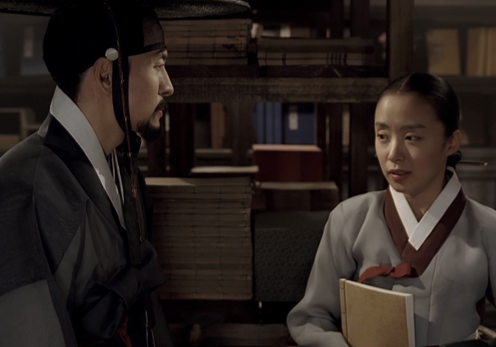 O Jo-won e a Sukbuin, que segura um livro, frente a frente numa sala com estantes
