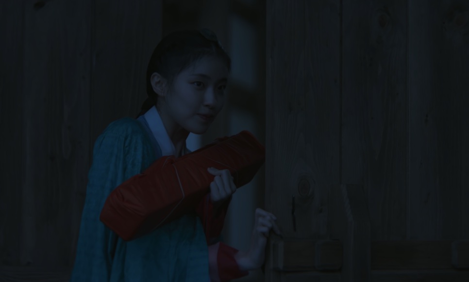 So-ok, de hanbok azul-claro, abraçada a um embrulho vermelho num canto escuro, em Escândalo e Intriga
