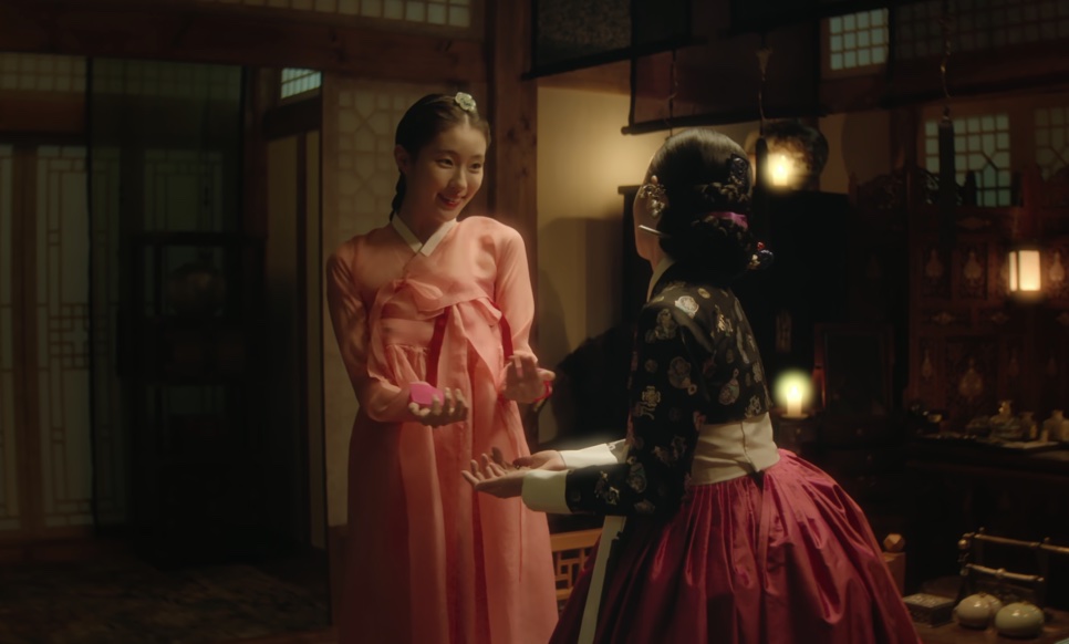 So-ok, de hanbok rosa, sorrindo diante de uma mulher de saia vermelha numa sala iluminada por lampiões, em Escândalo e Intriga