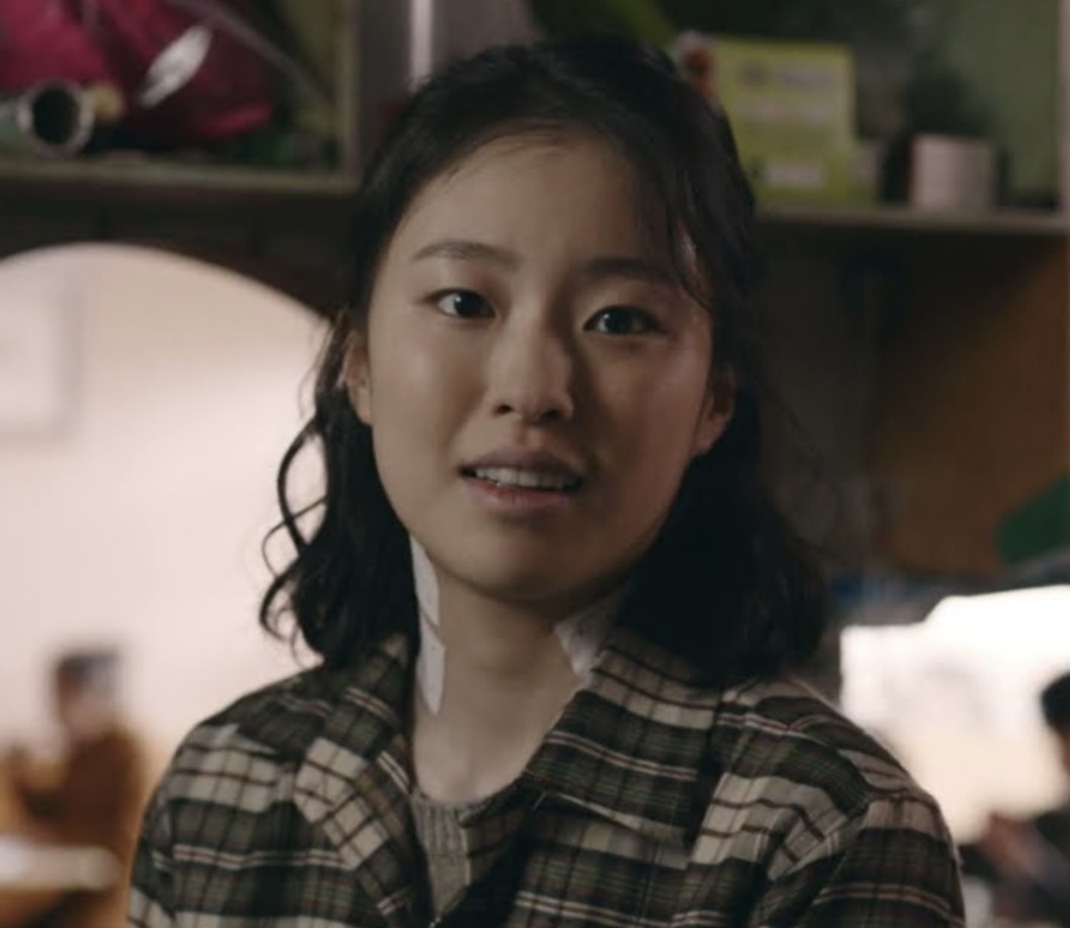 Shin Youn-ha como Lee Jeong-ok, de camisa xadrez e cabelo preso, numa cena da série Oasis