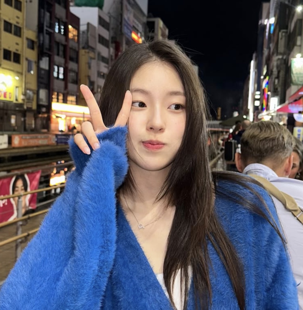 Shin Youn-ha de casaco azul peludo, fazendo sinal de V, numa rua iluminada por letreiros à noite