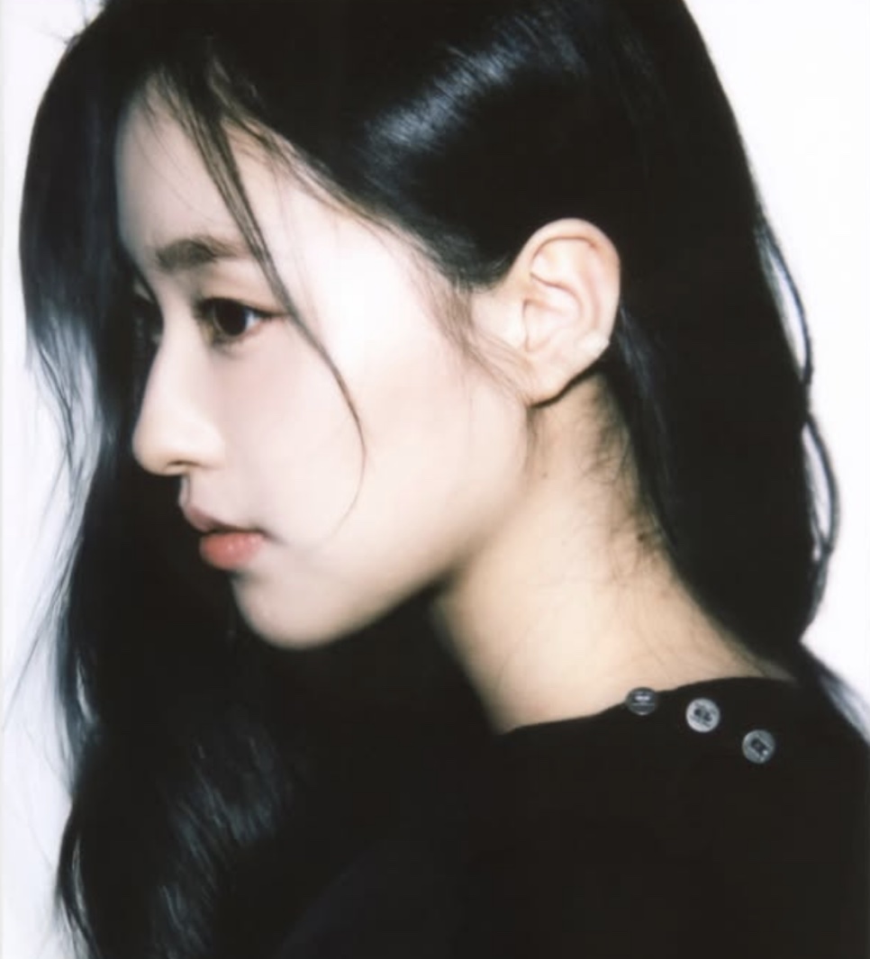 Retrato de perfil da Shin Youn-ha, de blusa preta e cabelo solto, sobre fundo claro