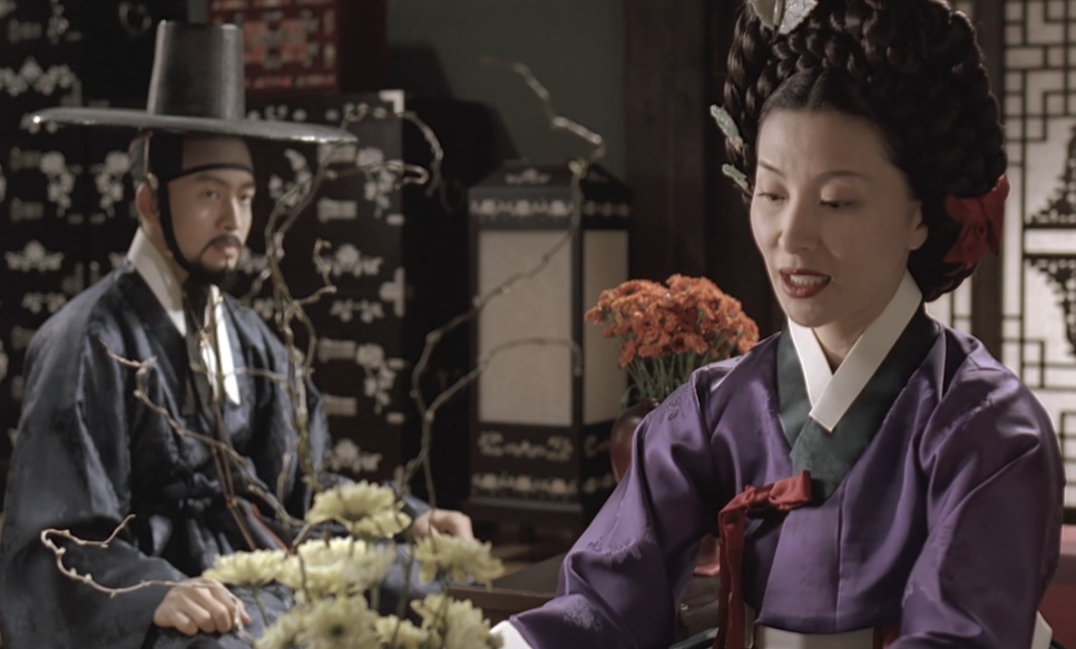 O Jo-won, de chapéu gat e roupa escura, e a Senhora Jo, de hanbok roxo, sentados diante de um arranjo de flores, em Untold Scandal