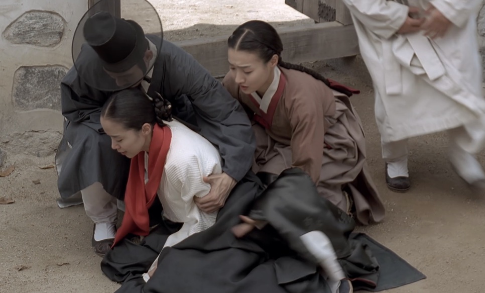 A Sukbuin desabando no chão, amparada por um homem de chapéu preto e por uma moça de hanbok, em Untold Scandal