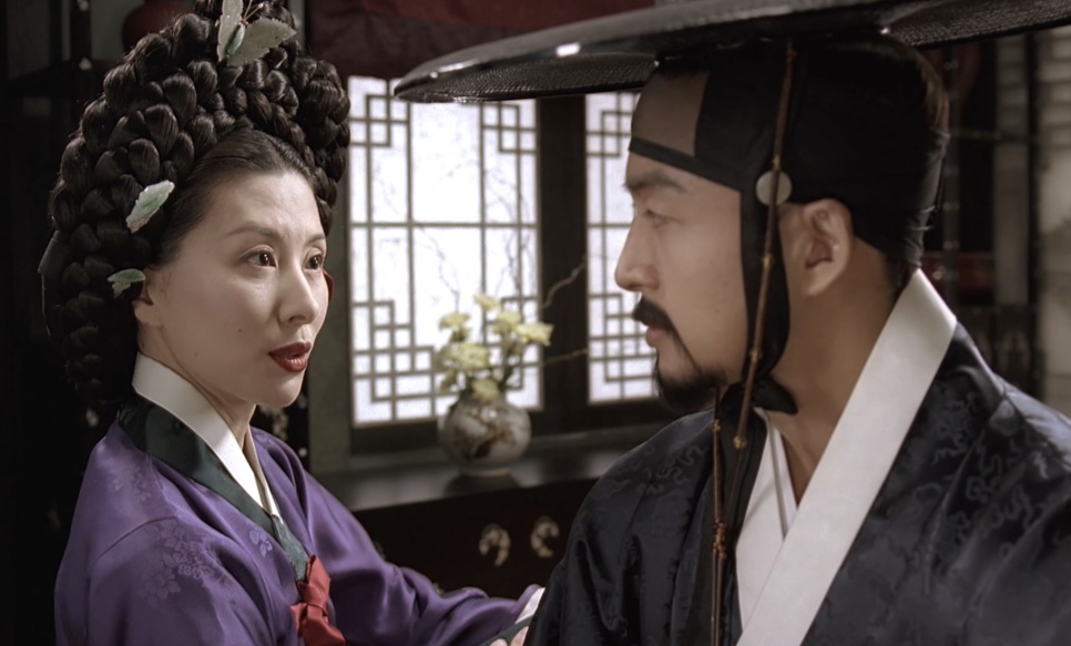 A Senhora Jo, de hanbok roxo e penteado alto, bem perto do Jo-won, de chapéu gat, numa sala com janelas de treliça, em Untold Scandal