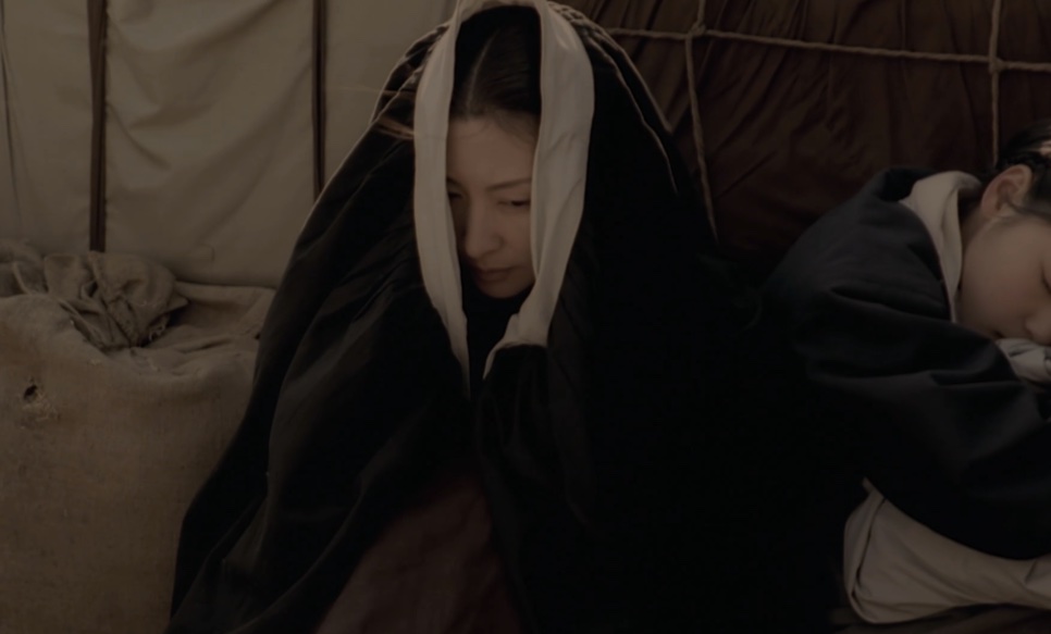 A Senhora Jo encolhida sob um manto escuro, com um pano claro na cabeça, ao lado de uma moça dormindo, em Untold Scandal