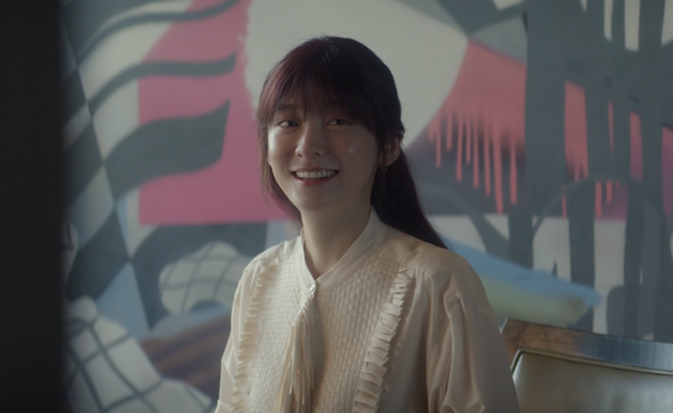 Cheon Sang-yeon (Park Ji-hyun) sorrindo de blusa clara, na frente de um painel colorido