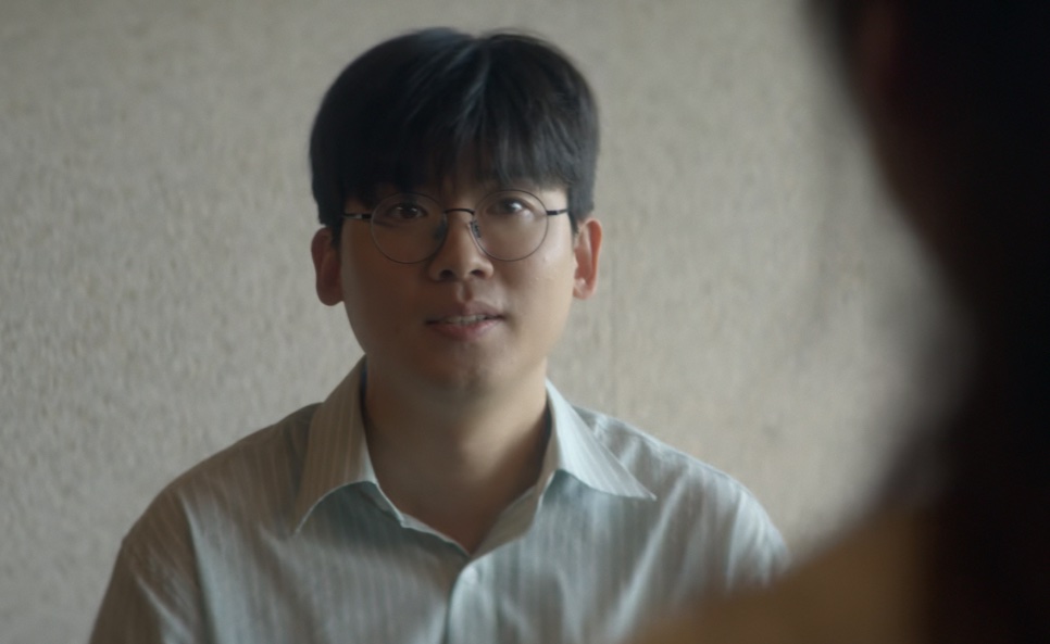 Han Jae-jun de óculos e camisa listrada, olhando para a pessoa sentada à sua frente