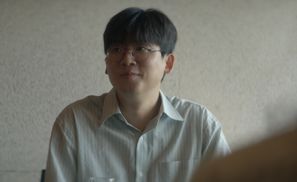 Han Jae-jun sentado à mesa, com um sorriso leve, em cena de Eu, Você e Toda Uma Vida