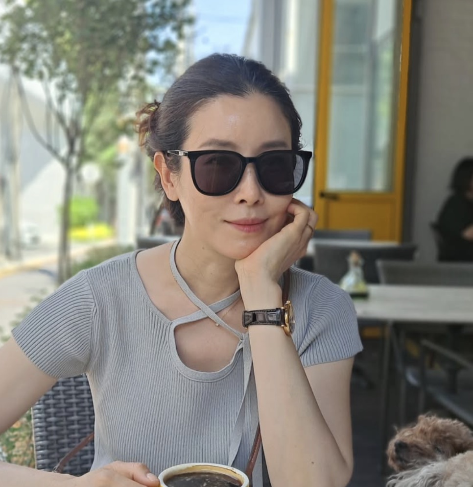 Lee Tae-ran de óculos escuros e blusa cinza, sentada numa mesa de cafeteria ao ar livre